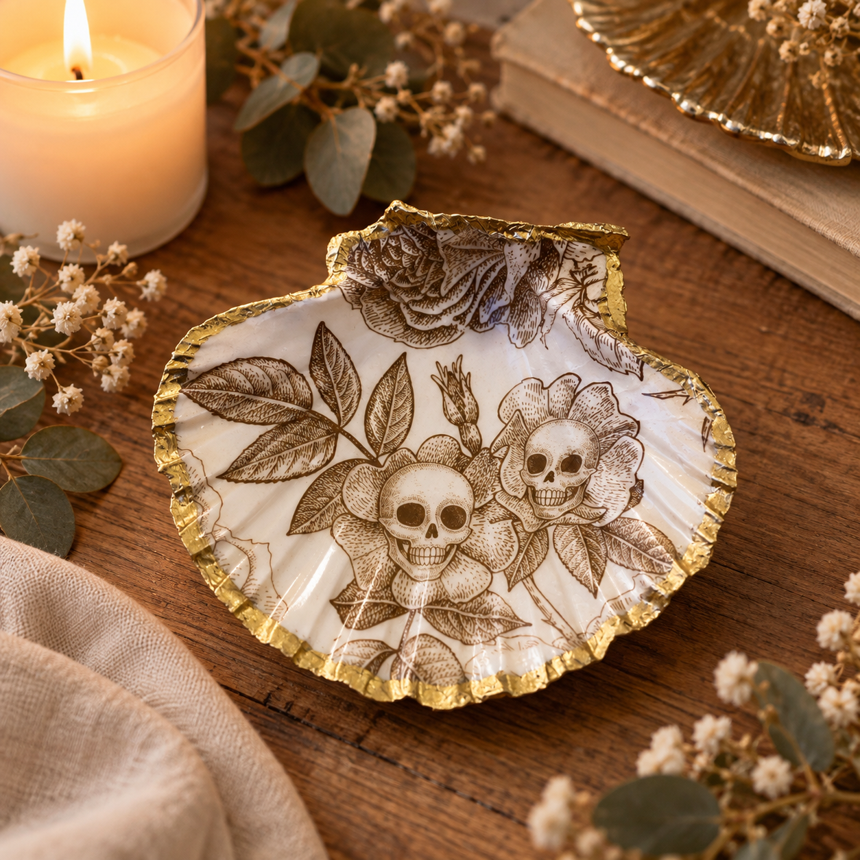 Coquille Saint-Jacques décorée de crânes et de roses gravés en sépia, bordure dorée à la feuille, posée sur une table en bois.